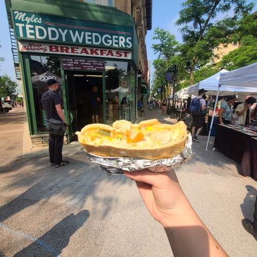 A hand holds a loaded breakfast sandwich in front of Teddy Wedgers restaurant, with people and market stalls in the background.