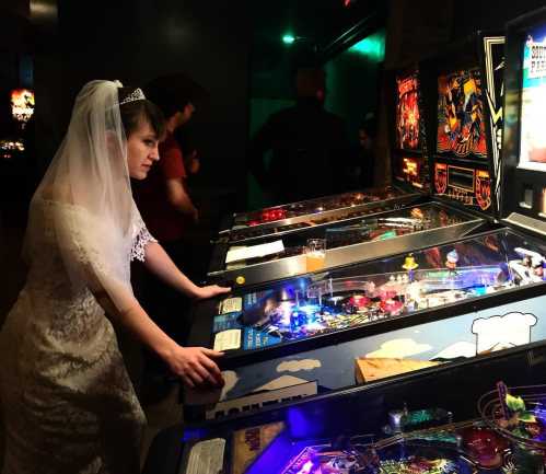 A bride in a wedding dress and veil plays pinball at an arcade, focused on the game with colorful lights around her.