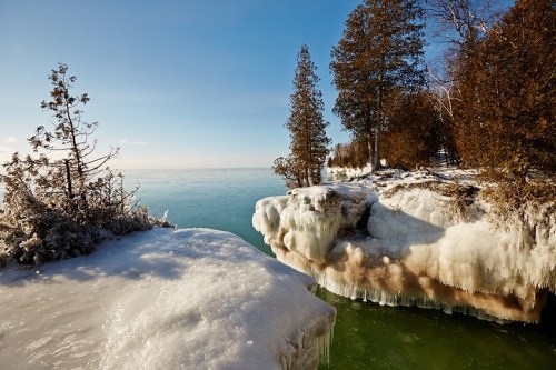 A serene winter landscape featuring icy cliffs and trees by a calm, frozen lake under a clear blue sky.
