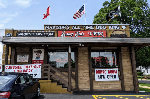 Exterior of Smoky Jon's BBQ restaurant with flags, signage, and a car parked outside. "Dining Room Now Open" displayed.