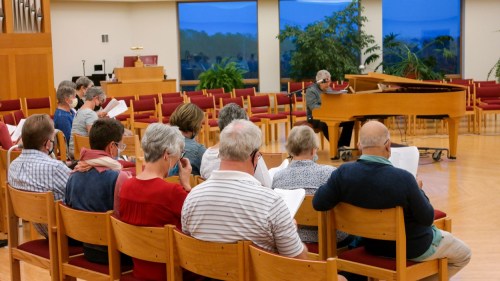 A group of people seated in a hall, singing along as a pianist plays at the front.
