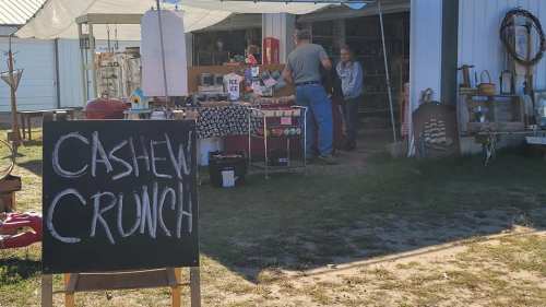 A market stall with a sign reading "Cashew Crunch," featuring various goods and two people browsing.