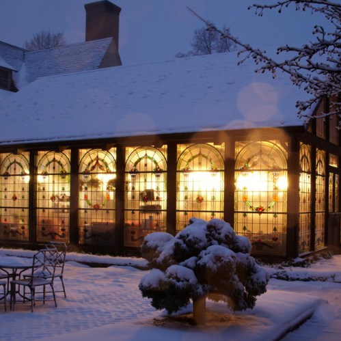 A cozy glass conservatory illuminated at dusk, surrounded by snow-covered trees and a winter landscape.