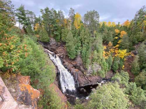 A scenic waterfall cascades down rocky cliffs, surrounded by lush green trees and autumn foliage.