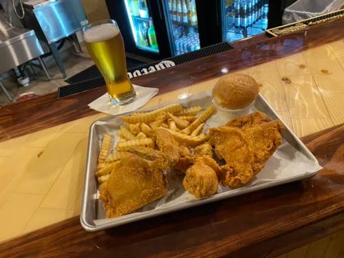 A tray with fried chicken, crinkle-cut fries, a bun, and a glass of beer on a bar counter.