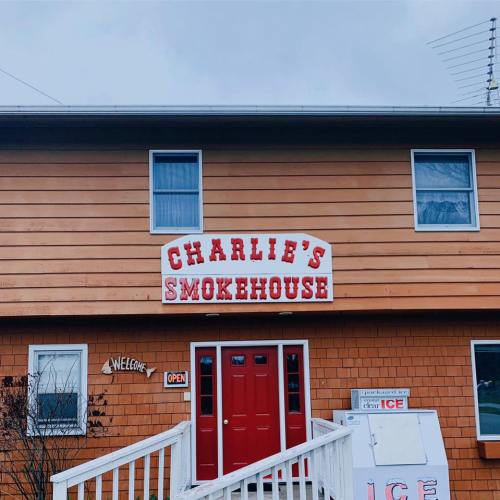 A wooden building with a sign reading "Charlie's Smokehouse" and a red door, featuring an open sign and ice display.