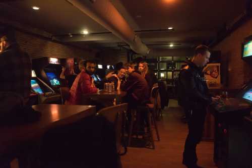 A dimly lit arcade bar with people socializing, playing games, and enjoying drinks at the counter and arcade machines.