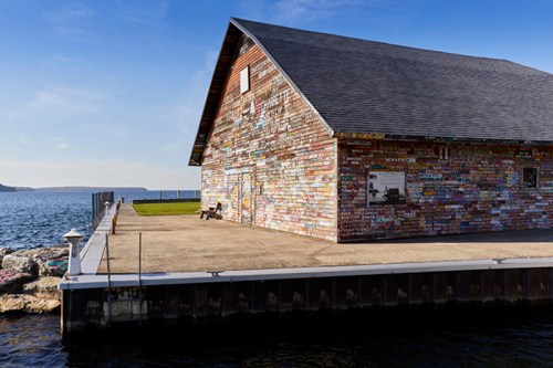 A weathered wooden building with colorful graffiti sits by the water, with a clear blue sky above.