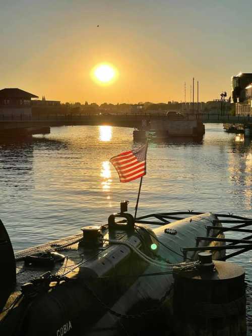 A sunset over a calm waterway, featuring a submarine with an American flag in the foreground.