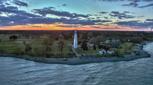 Aerial view of a lighthouse at sunset, surrounded by trees and a calm lake, with colorful clouds in the sky.