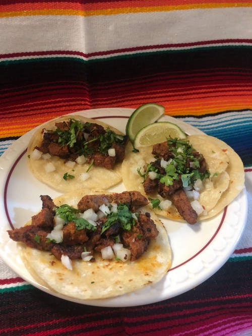Three tacos filled with seasoned meat, topped with onions and cilantro, served with lime wedges on a colorful striped background.