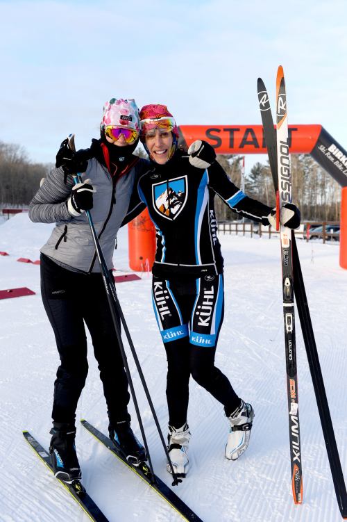 Two women in ski gear pose together with skis, smiling at the camera near a start line banner in a snowy landscape.