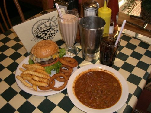 A plate with a burger, fries, onion rings, and a bowl of chili, accompanied by milkshakes and soda on a checkered table.