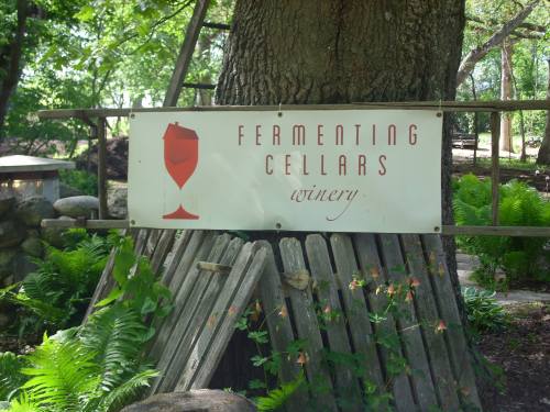 Sign for "Fermenting Cellars Winery" featuring a wine glass graphic, surrounded by greenery and wooden structures.