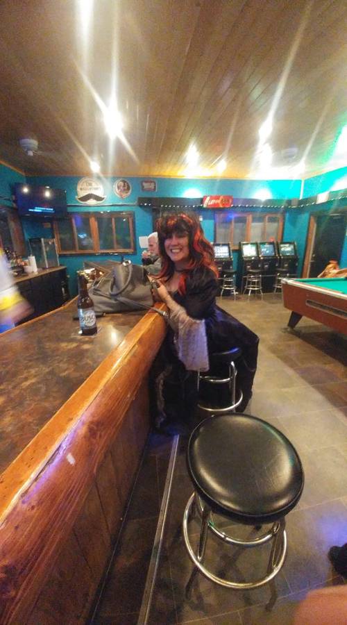 A woman with red hair and a black outfit smiles while sitting at a bar, holding a drink and a small stuffed animal.