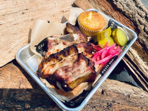 A metal tray with grilled ribs, pickles, pink onions, a cornbread muffin, and parchment paper, resting on wooden logs.