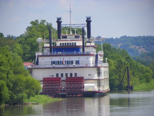 A large riverboat with two smokestacks docked along a lush green riverbank under a clear blue sky.