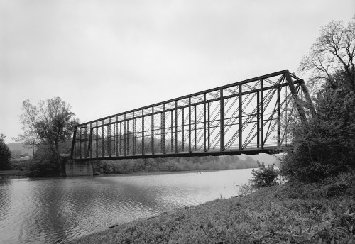 A black and white image of a metal truss bridge spanning a calm river, surrounded by trees and a cloudy sky.