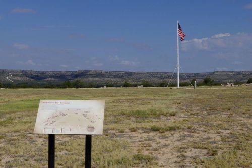 A grassy field with a sign and a tall flagpole displaying the American flag against a backdrop of distant hills.