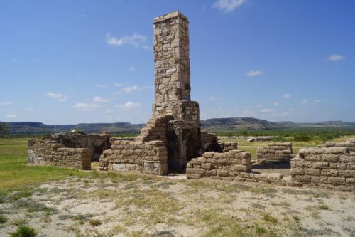 Ruins of a stone structure with a tall chimney, set against a clear blue sky and distant hills.