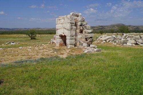 A weathered stone ruin stands in a grassy field, surrounded by rolling hills under a blue sky with scattered clouds.
