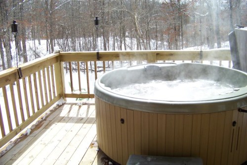 A steaming hot tub on a wooden deck surrounded by a snowy forest landscape.