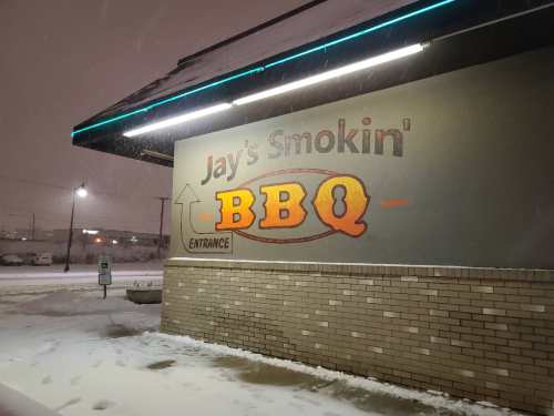 Sign for "Jay's Smokin' BBQ" illuminated at night, with snow covering the ground and a snowy atmosphere.