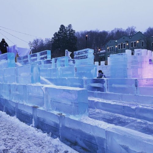 A winter scene featuring a large ice sculpture maze with blue-tinted ice blocks and people exploring.