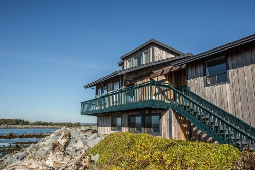 A modern wooden house with a green staircase, overlooking a rocky shoreline and clear blue sky.
