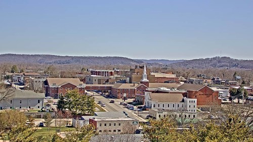 A panoramic view of a small town with buildings, a church steeple, and rolling hills in the background under a clear blue sky.