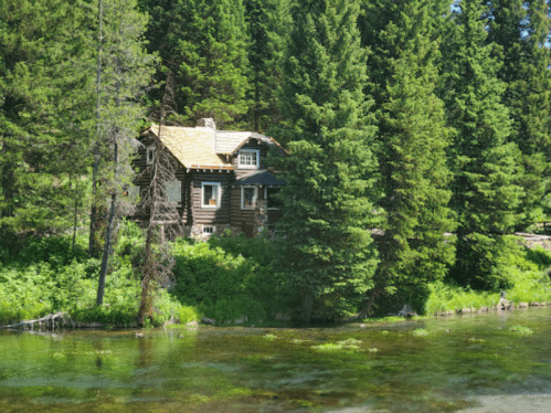 A rustic log cabin surrounded by tall green trees, next to a clear, calm river.
