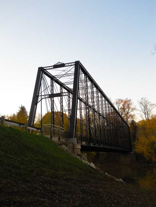 A metal truss bridge spans a river, surrounded by trees with autumn foliage under a clear sky.