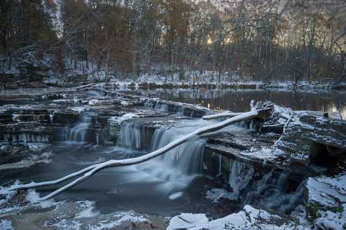 A serene winter scene featuring a frozen waterfall, icy rocks, and a fallen tree, surrounded by a tranquil forest.