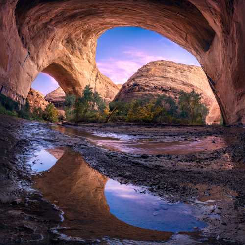 A serene canyon scene with a large arch, reflecting water, and colorful sky at sunset. Lush greenery surrounds the area.