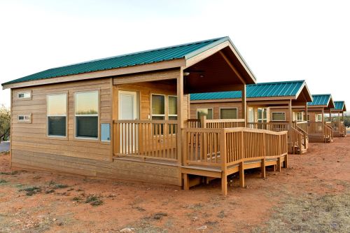 Four wooden cabins with green roofs and porches, set in a sandy outdoor area.