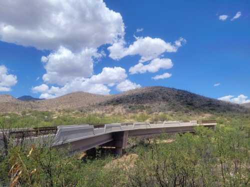 A concrete bridge spans a dry landscape with rolling hills and a blue sky dotted with fluffy white clouds.