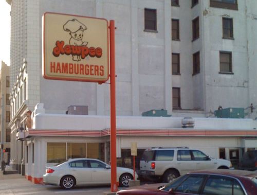 Sign for "Newpee Hamburgers" in front of a building, with parked cars in the foreground.