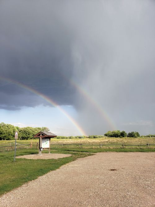 A double rainbow arcs over a grassy field under dark, stormy clouds, with a gravel path in the foreground.