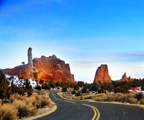 A winding road leads through red rock formations under a blue sky, with snow-dusted ground and scattered trees.