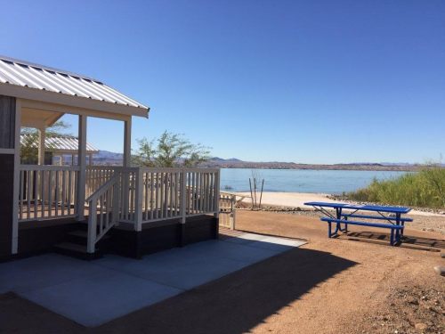A lakeside view featuring a small cabin, picnic table, and clear blue water under a bright sky.