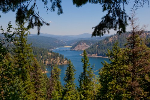 A serene view of a blue lake surrounded by lush green trees and mountains under a clear blue sky.