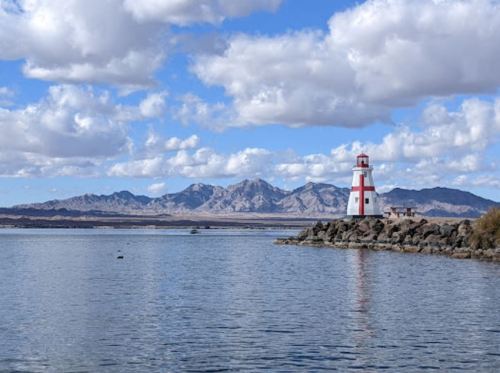 A scenic view of a lighthouse by the water, with mountains and fluffy clouds in the background.