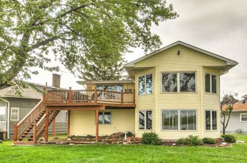 A modern yellow house with a large deck, surrounded by green grass and trees, under a cloudy sky.