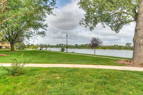 A serene park scene featuring a grassy path along a calm lake, surrounded by trees and cloudy skies.