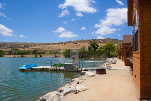A serene lakeside view with a wooden cabin, dock, and paddle boats under a blue sky with fluffy clouds.