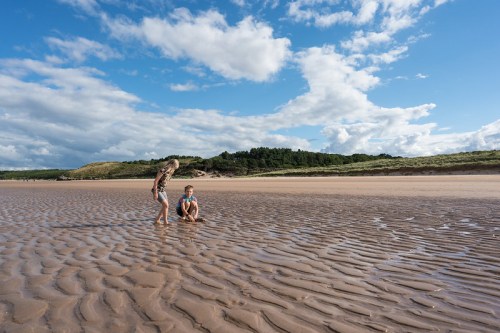 Two children playing on a sandy beach with ripples, under a blue sky with fluffy clouds.