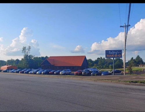 A barbecue restaurant with a red roof and multiple parked cars in front, set against a blue sky with fluffy clouds.