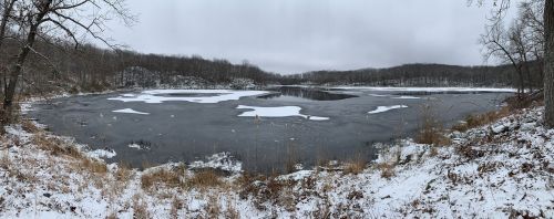 A snowy landscape featuring a partially frozen lake surrounded by trees and hills under a cloudy sky.