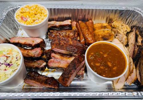 A tray of assorted barbecue meats, including ribs, sausage, and chicken, with sides of coleslaw, baked beans, and popcorn.