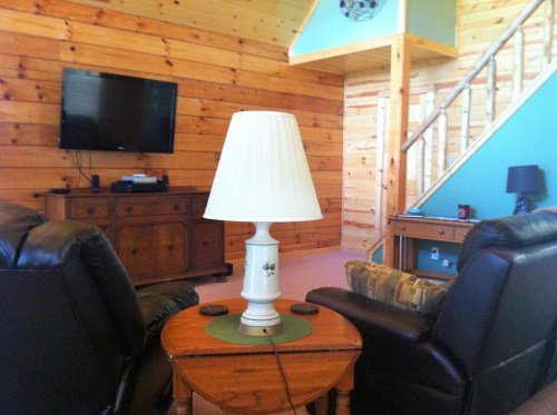Cozy living room with wooden walls, a TV, two leather chairs, a lamp, and a staircase in the background.